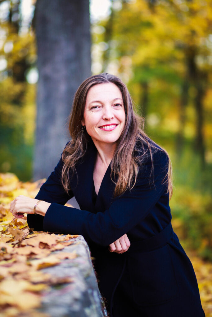 Astrid Bohm lehnt lächelnd an einem großen Stein in einem herbstlichen Wald und trägt einen dunklen Mantel